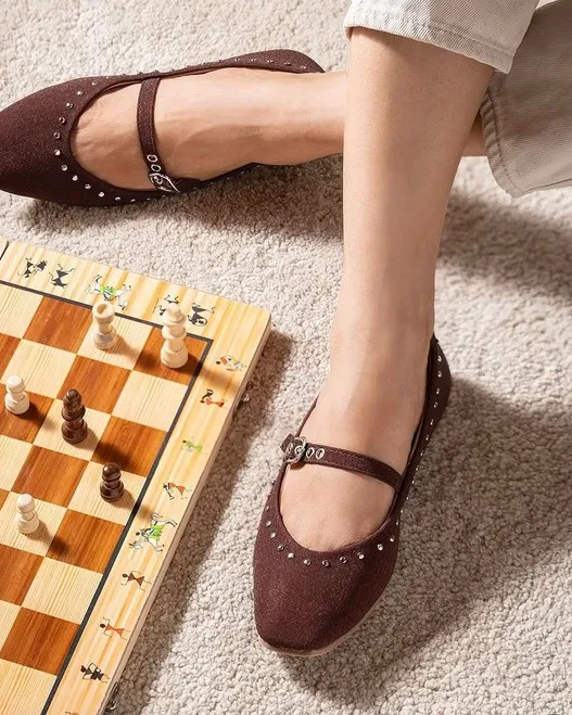 Studded brown mary-jane shoes worn beside a wooden chessboard on a cream carpet