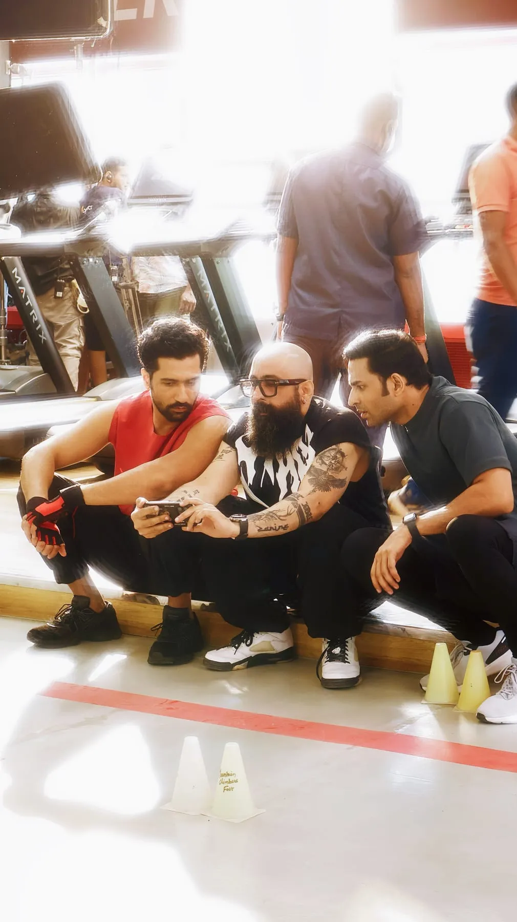 Three men crouched on a gym floor between treadmills, looking at a phone during a break in the shoot
