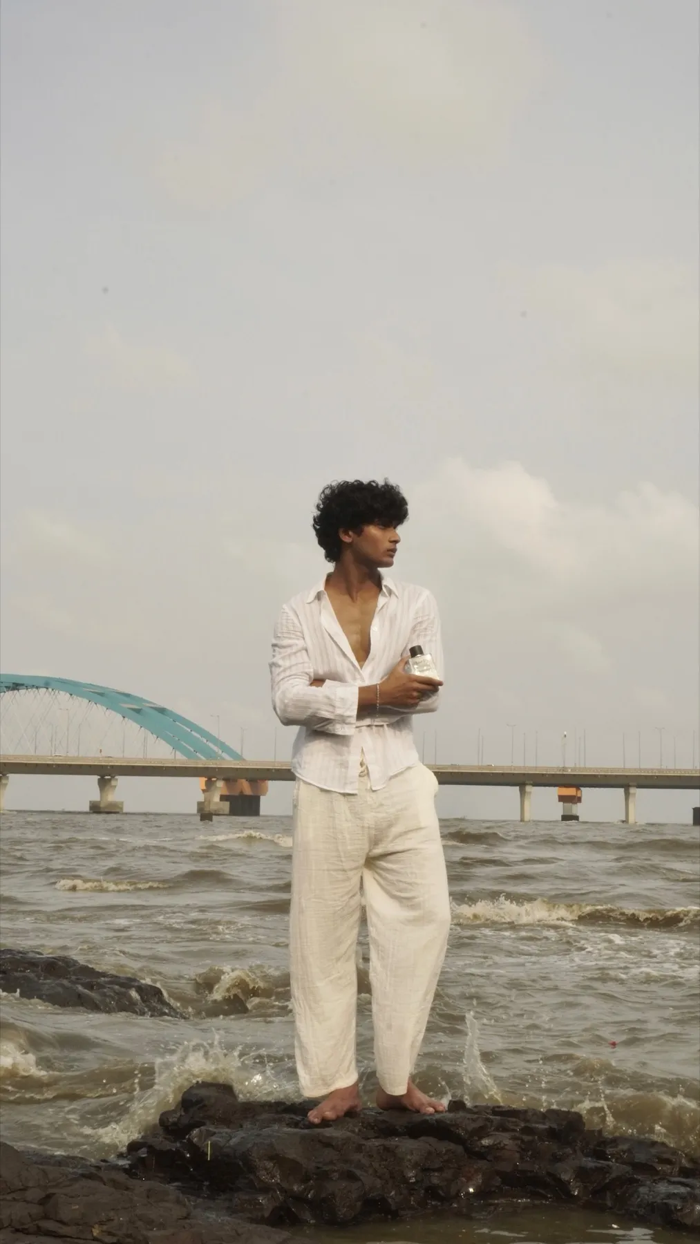 Man in white linen holding a Sanso bottle on sea rocks, with the Bandra–Worli sea link behind